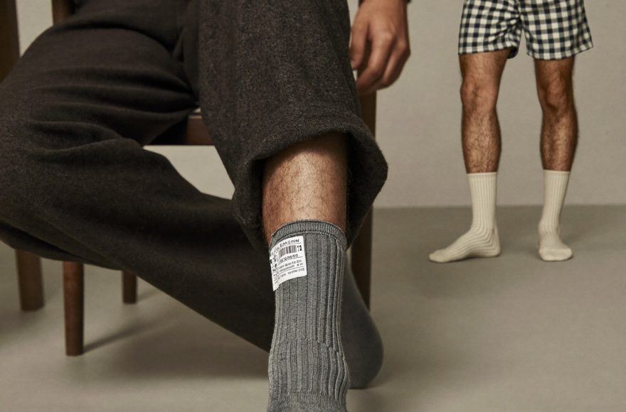 A seated man's crossed leg showing several inches of bare shin between the top of a grey crew sock and a charcoal trouser hem