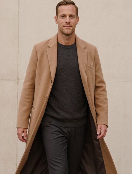 Tall man walking in an open camel wool overcoat that finishes below the knee over a charcoal sweater and trousers