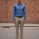 A tall man standing against a brick wall in a blue long-sleeve shirt tucked into stone-coloured chinos with white sneakers.