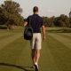 A tall man walking a mown golf course fairway carrying a golf bag, wearing stone-grey golf shorts that finish above the knee and a navy polo shirt.
